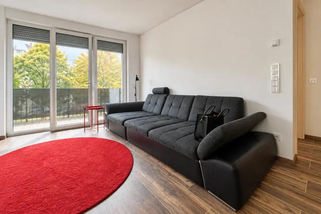 Helles Wohnzimmer mit großer Fensterfront, Balkonzugang, dunkelgrauem Sofa und rotem Teppich